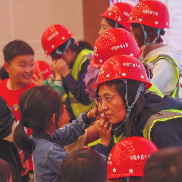 六一 5月29日，在中鐵十局一公司安全講解員的帶領下，來自公司近二十組家庭的萌娃們化身小小“安全員”，與父母共同參與了在遙墻機場項目部開展的“萌娃進工地，童聲囑安全”親子活動。圖為孩子們為爸爸媽媽佩戴安全帽。